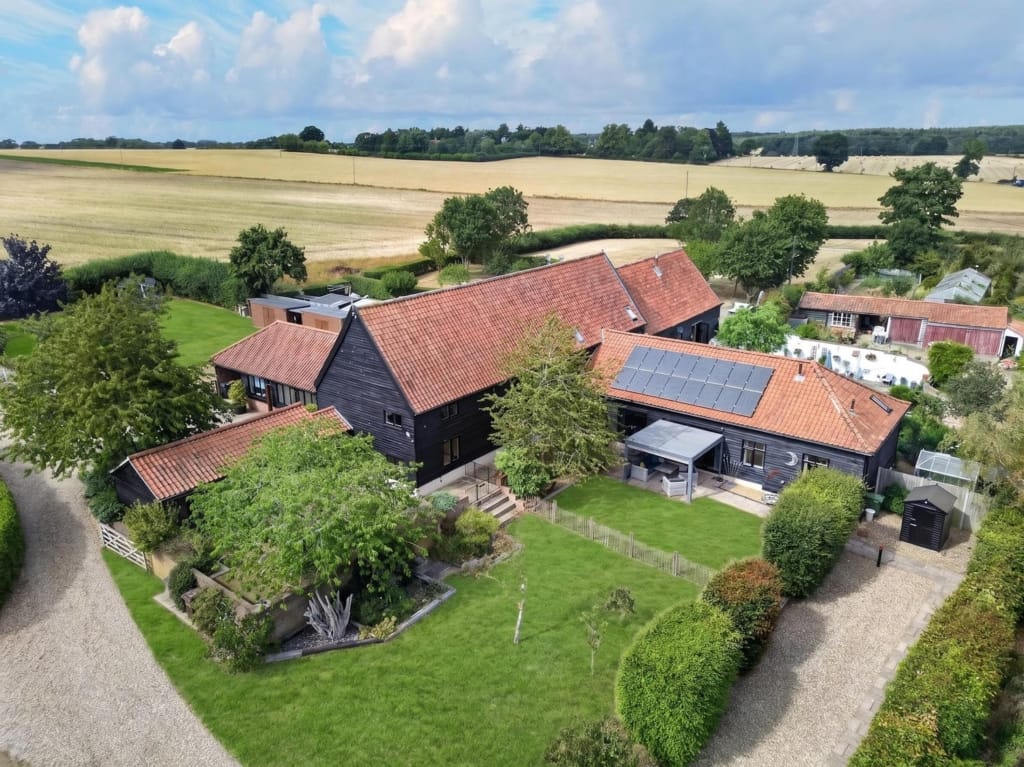 Aerial view of The Tithe Barn Sisland showing dark-clad barn complex, terracotta roofs, solar panels, lawned gardens, and surrounding Norfolk arable fields