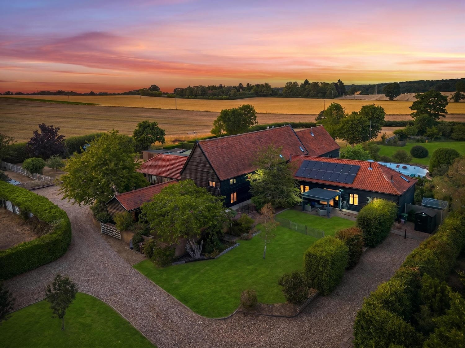 Aerial drone view of The Tithe Barn at sunset, dark timber barn buildings with terracotta roofs, lawned grounds, gravel driveway, and open Norfolk arable fields beyond