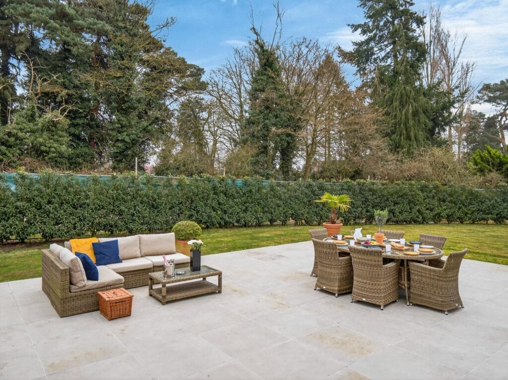 Spacious paved patio with outdoor dining and seating area overlooking a large private garden at a modern luxury home.