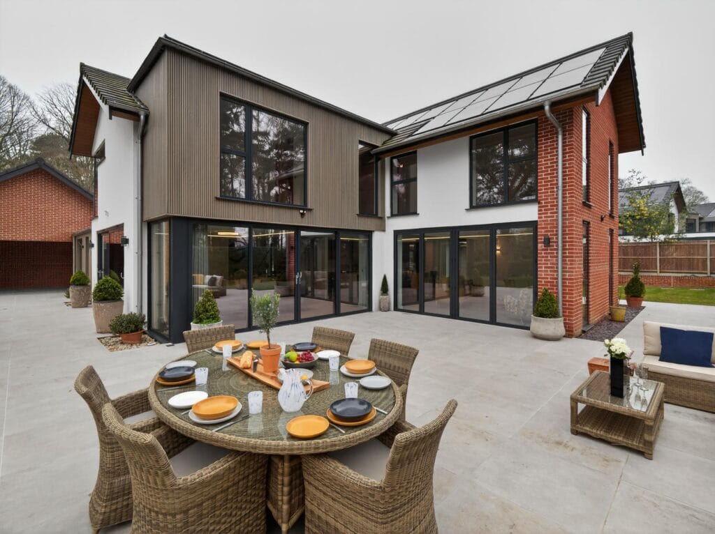 Large patio terrace with outdoor dining area overlooking the modern home with floor-to-ceiling glazing and landscaped garden.