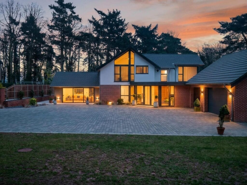 Luxury contemporary detached house with large driveway, floor-to-ceiling windows and evening lighting.