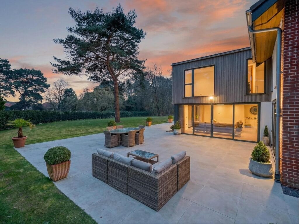 Luxury home patio and garden at dusk with outdoor seating and floor-to-ceiling glazing illuminating the interior.
