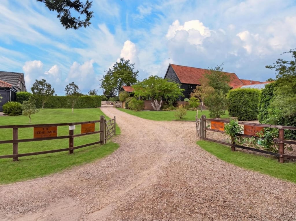 Gated gravel entrance drive leading to The Tithe Barn Sisland with black-clad barn buildings and red pantile roofs beyond mature hedging, Sisland, Norfolk