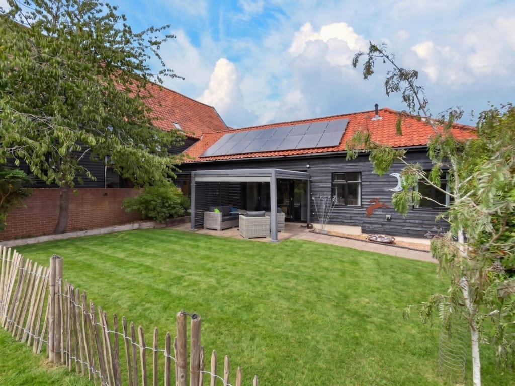 Rear garden of The Tithe Barn Sisland showing dark-clad barn, solar panels, pergola with rattan seating, and lawned grounds
