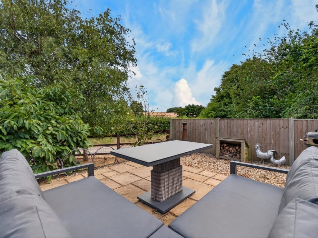 Paved terrace seating area with rattan table, log store, and mature tree surround at The Tithe Barn, Sisland, Norfolk