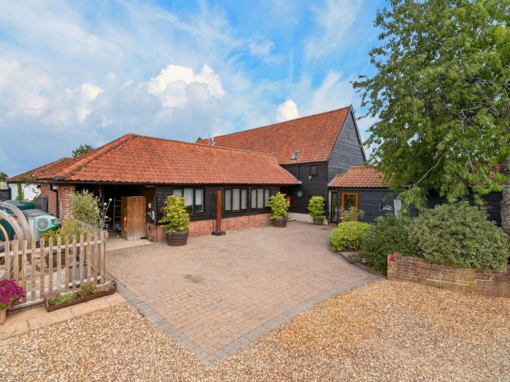 Exterior of The Tithe Barn Sisland showing black timber cladding, red pantile roofs, and block-paved courtyard with gravel forecourt, Norfolk
