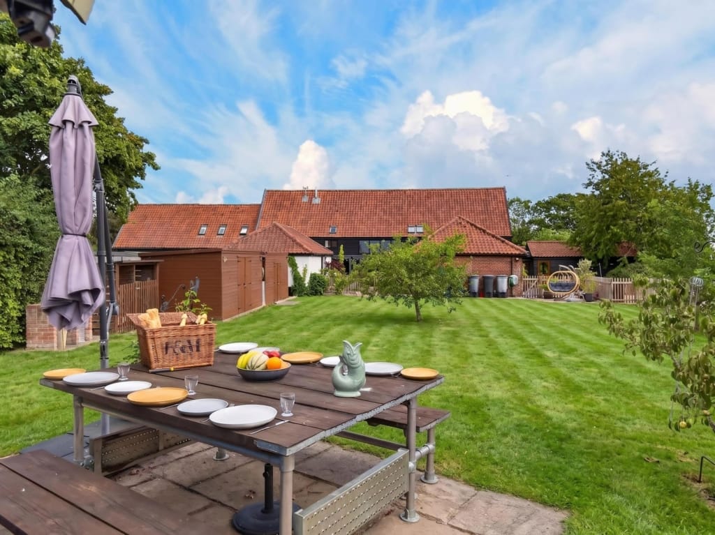 Gardens and grounds at The Tithe Barn Sisland Norfolk, laid dining table on stone terrace, striped lawn, barn complex beyond