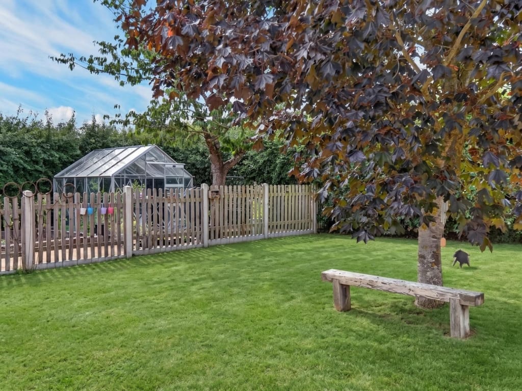 Gardens at The Tithe Barn Sisland showing lawn, timber bench beneath copper-leafed tree, picket fence, and aluminium greenhouse beyond