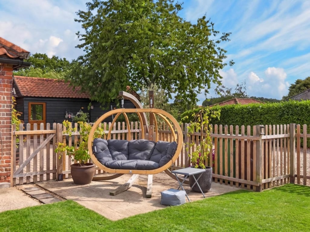 Garden seating area with hanging egg chair on paved terrace, picket fence and outbuilding beyond, The Tithe Barn, Sisland, Norfolk