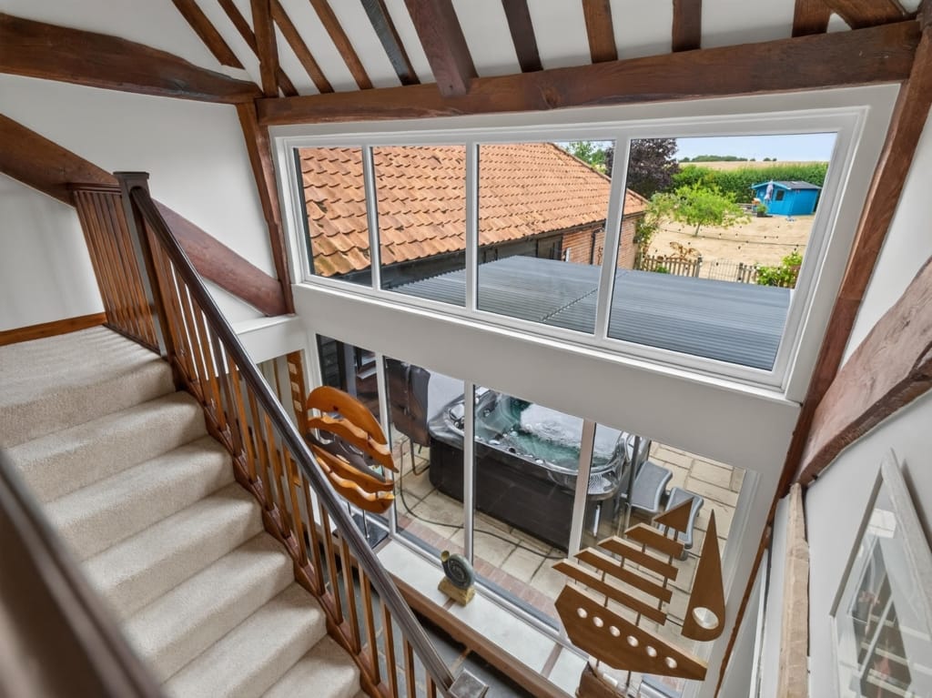 Staircase landing with oak balustrade, exposed timber beams, and large window overlooking courtyard hot tub and outbuildings at The Tithe Barn, Sisland, Norfolk