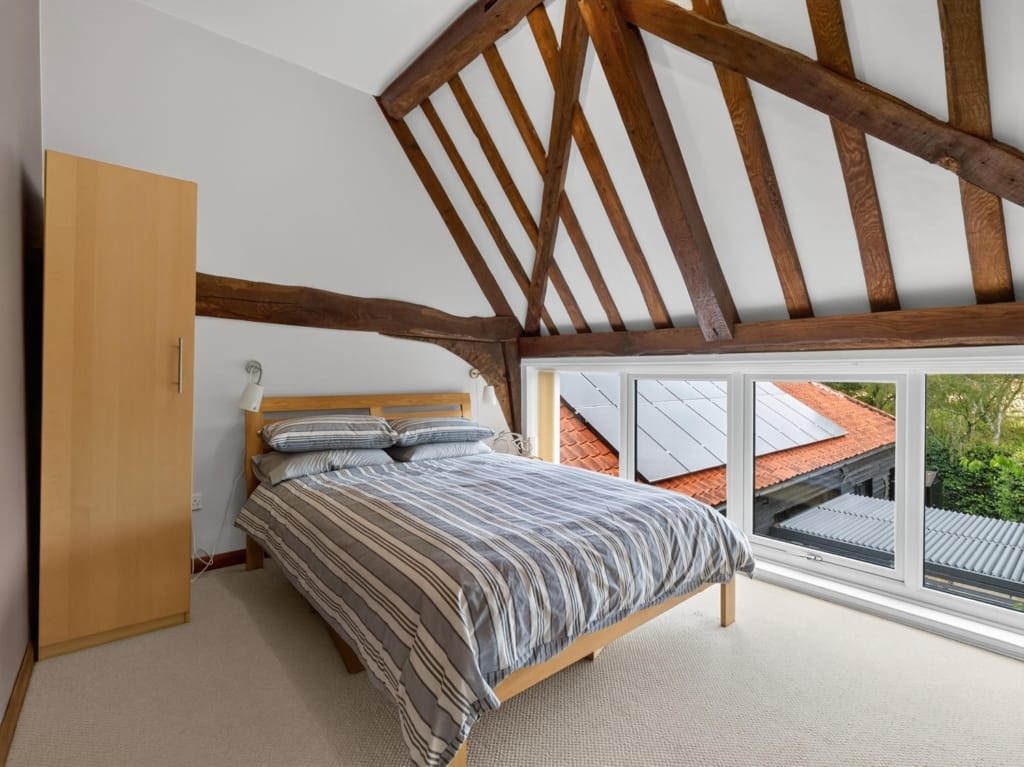 Bedroom with exposed oak truss framework, double bed, and wide window overlooking barn rooftops at The Tithe Barn, Sisland, Norfolk