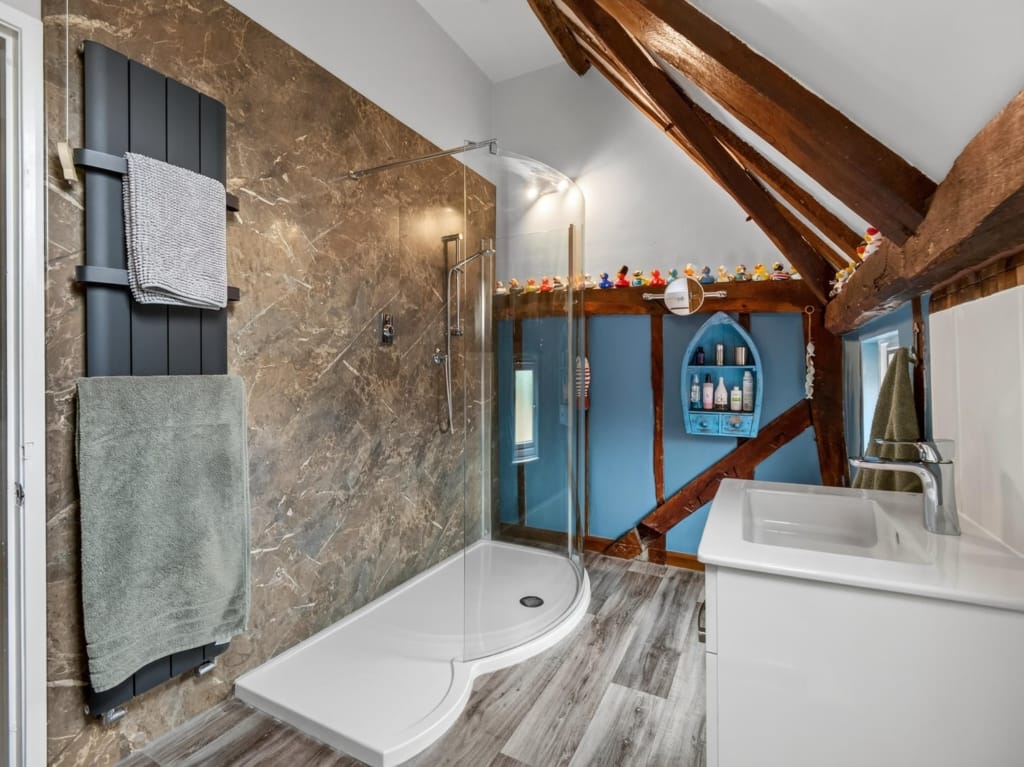 Family bathroom with curved glass shower enclosure, marble-effect tiled wall, exposed timber trusses, and blue-painted alcove shelving at The Tithe Barn, Sisland, Norfolk