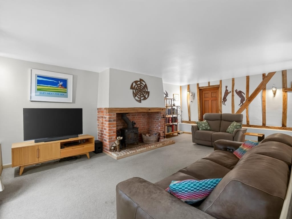 Sitting room with brick inglenook fireplace, wood-burning stove, exposed timber frame wall, and leather sofas at The Tithe Barn, Sisland, Norfolk