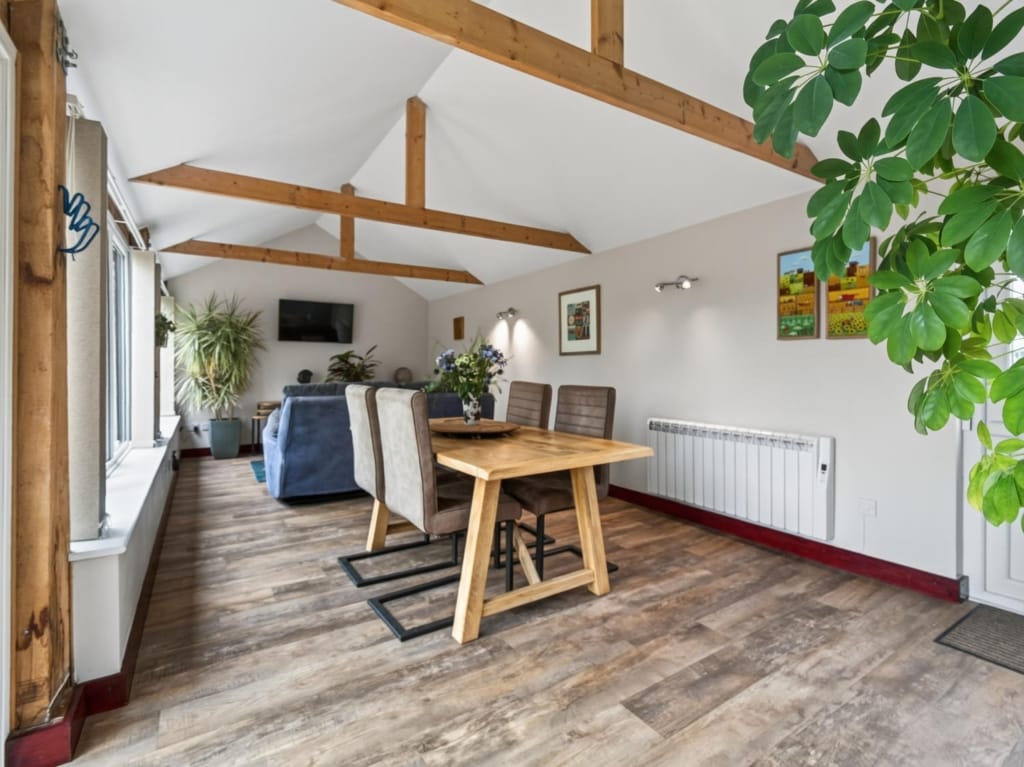 Open-plan living and dining area with vaulted oak beams, oak dining table, and wood-effect floor in one of the holiday barns at The Tithe Barn, Sisland, Norfolk