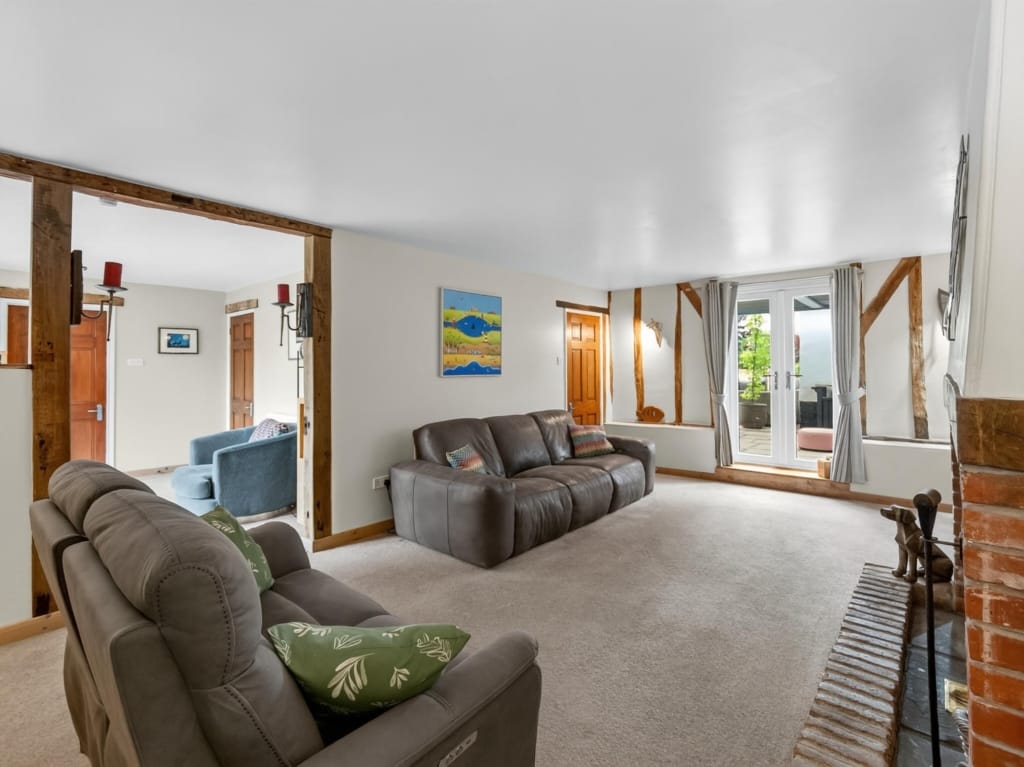 Sitting room with exposed timber frame, leather sofas, brick fireplace and French doors to terrace, The Tithe Barn, Sisland, Norfolk