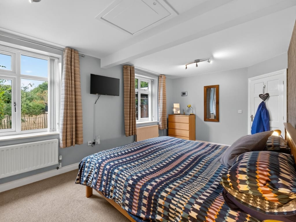Double bedroom with patterned bedspread, wall-mounted television, and two garden-facing windows at The Tithe Barn, Sisland, Norfolk