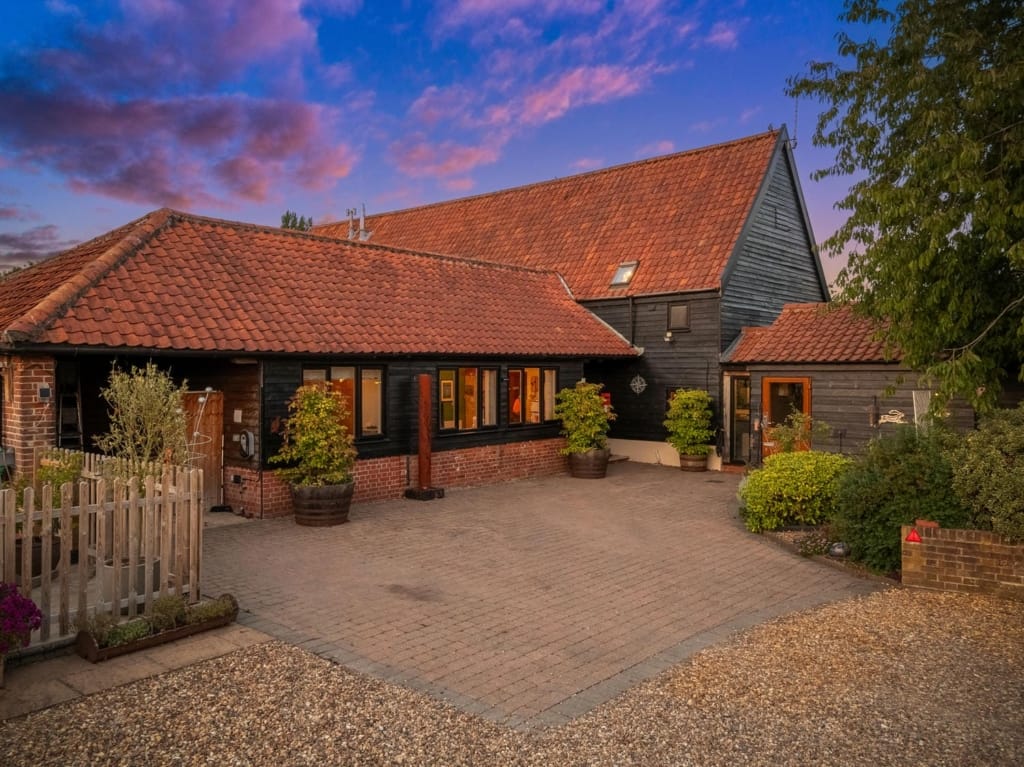 Exterior of The Tithe Barn at dusk, black timber cladding, red pantile roofs, and block-paved courtyard, Sisland, Norfolk