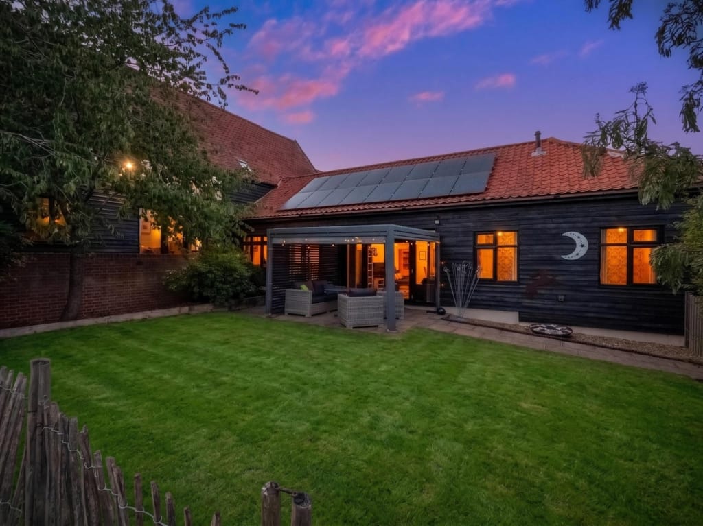Rear garden at dusk, black-clad barn with pergola seating area, solar panels, and glowing windows, The Tithe Barn, Sisland, Norfolk