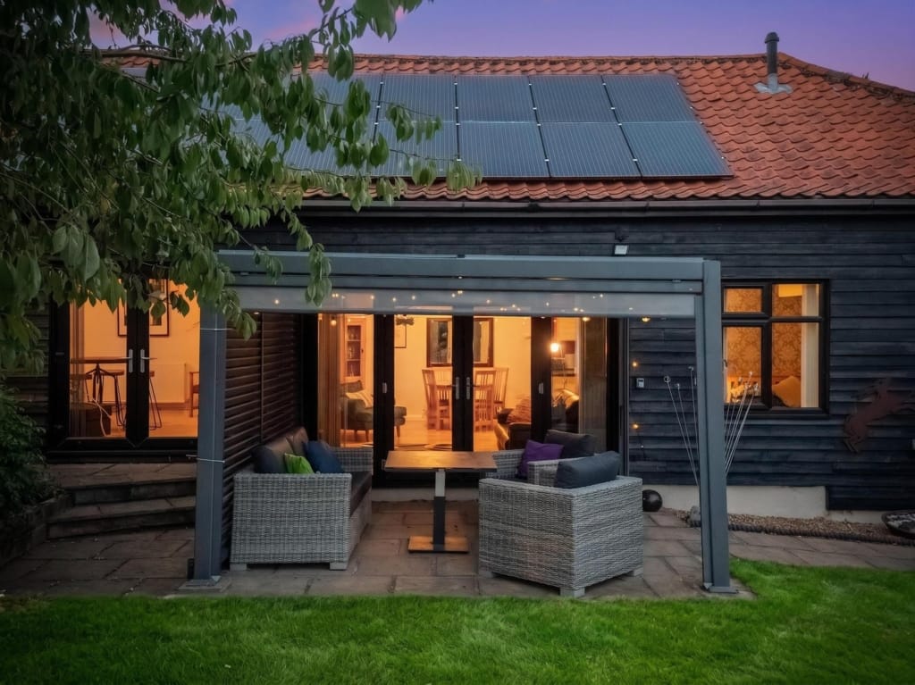Gardens and grounds at dusk, glazed pergola with rattan seating, bi-fold doors open to lit interior, solar panels on pantile roof, The Tithe Barn, Sisland, Norfolk