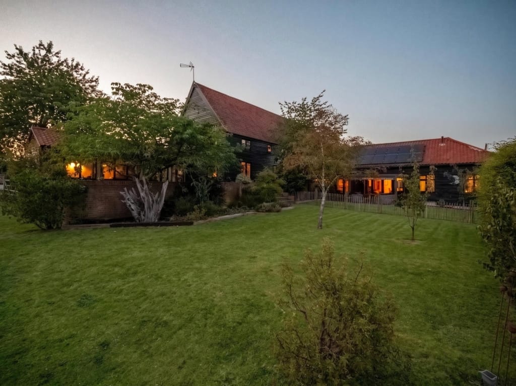 Dusk exterior of The Tithe Barn Sisland showing black-clad barn buildings, glowing lit windows, red pantile roofs, and wide lawn, Norfolk